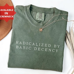 Peut inclure: Un t-shirt vert olive avec l'inscription blanche "RADICALIZED BY BASIC DECENCY". Un collier doré avec un pendentif est posé sur le t-shirt. Un chapeau marron et une feuille décorative sont également dans l'image.