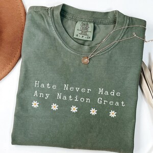 Può includere: Una maglietta verde oliva con la scritta "Hate Never Made Any Nation Great" e grafiche di margherite. Una collana d'oro con un ciondolo è appoggiata sulla maglietta. Un cappello marrone e piante secche sono sullo sfondo.