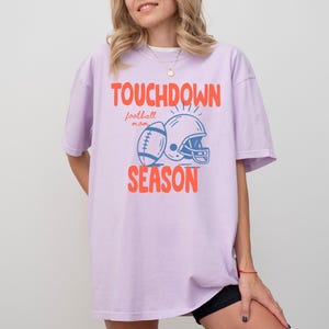 Puede incluir: Camiseta lila claro con las palabras "TOUCHDOWN SEASON" en naranja, junto con un bal&oacute;n de f&uacute;tbol y un casco azules. La camiseta es de estilo casual, ideal para los aficionados al f&uacute;tbol.