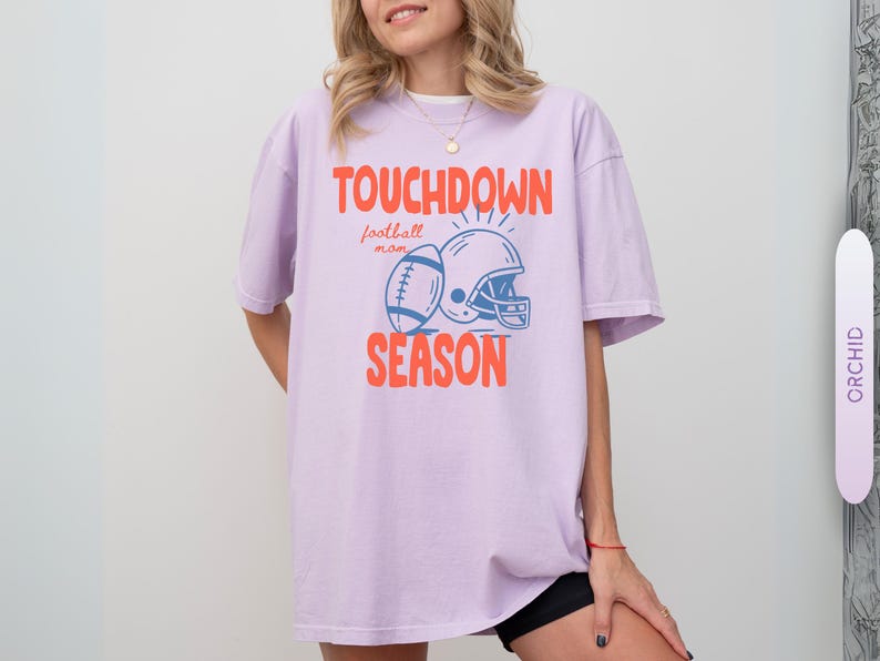 Puede incluir: Camiseta lila claro con las palabras "TOUCHDOWN SEASON" en naranja, junto con un bal&oacute;n de f&uacute;tbol y un casco azules. La camiseta es de estilo casual, ideal para los aficionados al f&uacute;tbol.