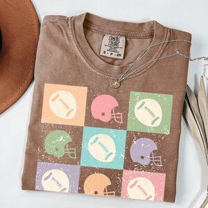 Peut inclure: T-shirt couleur expresso avec un motif en grille de ballons et de casques de football américain aux couleurs pastel. Le t-shirt a un col rond et des manches courtes. Un collier argenté avec un petit pendentif repose sur le t-shirt.