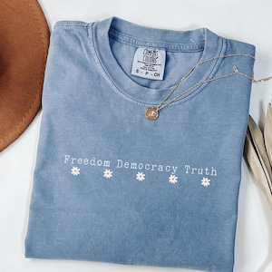 Peut inclure: Un t-shirt bleu jean avec les mots "Freedom Democracy Truth" et quatre motifs de marguerites. Un collier en or avec un pendentif repose sur le t-shirt. Le t-shirt est étiqueté "Comfort Colors" et est de taille S-CH.