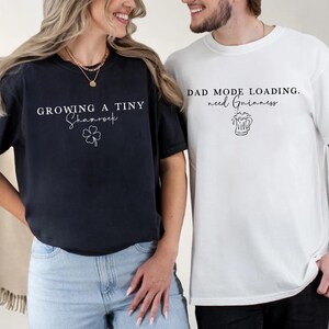 May include: Two t-shirts, one navy blue and one white, with text and graphics. The navy shirt says "Growing a Tiny Shamrock" with a shamrock graphic. The white shirt says "Dad Mode Loading. need Guinness" with a beer mug graphic.