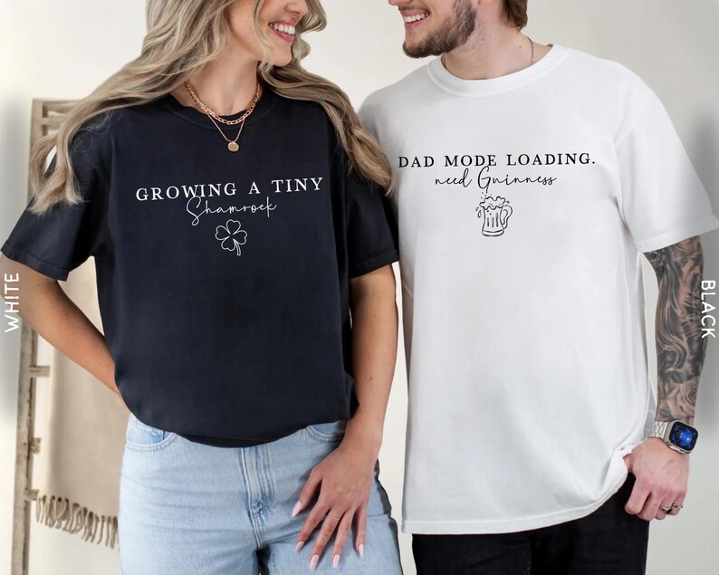 May include: Two t-shirts, one navy blue and one white, with text and graphics. The navy shirt says "Growing a Tiny Shamrock" with a shamrock graphic. The white shirt says "Dad Mode Loading. need Guinness" with a beer mug graphic.