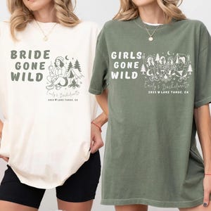 Puede incluir: Dos camisetas holgadas, una marfil y otra verde musgo, con texto gr&aacute;fico e ilustraciones. La camiseta marfil dice "BRIDE GONE WILD" con una escena de acampada. La camiseta verde dice "GIRLS GONE WILD" con una escena de acampada en grupo. Ambas camisetas incluyen "Emily's Bachelorette" y la ubicaci&oacute;n.