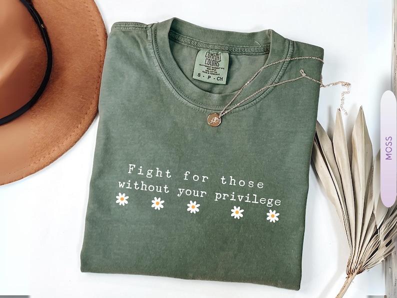 May include: Moss green t-shirt with the text "Fight for those without your privilege" and daisy graphics. A gold necklace and a brown hat are also visible. The shirt is folded on a white surface.