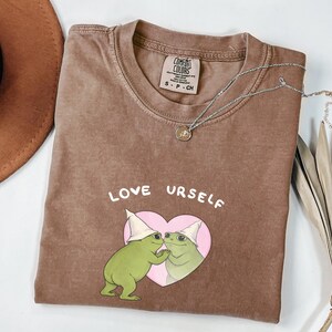 Pu&ograve; includere: T-shirt color espresso con le parole "LOVE URSELF" sopra un cuore rosa con due illustrazioni di rane verdi. La maglietta ha un girocollo e una piccola etichetta. Una collana d'argento con un ciondolo &egrave; sulla maglietta.