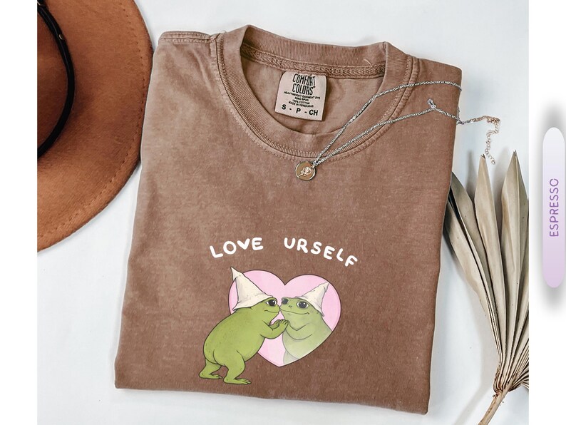 Pu&ograve; includere: T-shirt color espresso con le parole "LOVE URSELF" sopra un cuore rosa con due illustrazioni di rane verdi. La maglietta ha un girocollo e una piccola etichetta. Una collana d'argento con un ciondolo &egrave; sulla maglietta.