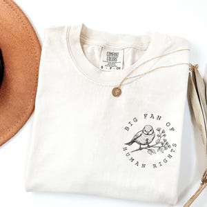 Peut inclure: T-shirt ivoire à col rond. Le t-shirt présente une illustration en noir et blanc d'un oiseau perché sur une branche, entourée du texte "BIG FAN OF HUMAN RIGHTS". Un collier en or avec un pendentif est drapé sur le t-shirt.