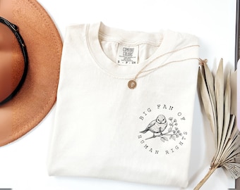 May include: An ivory short-sleeve t-shirt featuring a black ink illustration of a bird perched on a branch. The circular graphic includes the text "BIG FAN OF HUMAN RIGHTS" surrounding the bird. The shirt has a Comfort Colors brand label.