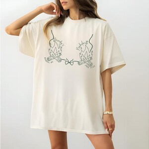 May include: Ivory-colored oversized t-shirt featuring a green graphic of two campfires with text. The text includes the words "Bachelorette" and "Hudson, NY". The shirt has short sleeves and a crew neck.