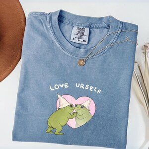 Pu&ograve; includere: Una maglietta blu Comfort Colors con la scritta "LOVE URSELF" e una grafica di due rane verdi in un cuore rosa. Sono visibili anche una collana d'oro e un cappello marrone.