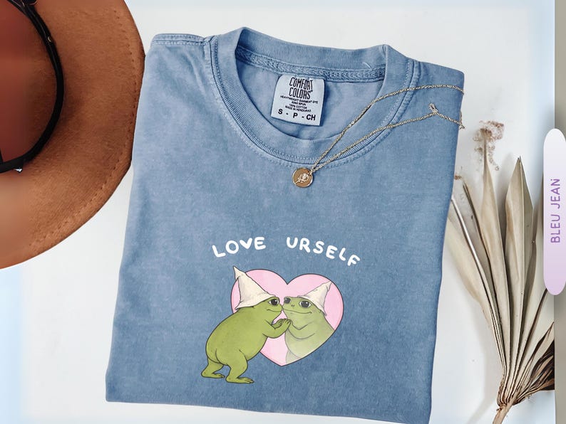 Pu&ograve; includere: Una maglietta blu Comfort Colors con la scritta "LOVE URSELF" e una grafica di due rane verdi in un cuore rosa. Sono visibili anche una collana d'oro e un cappello marrone.
