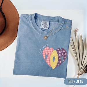 Può includere: Una t-shirt color blu jeans con scollo rotondo e una grafica sul davanti. La grafica presenta la parola MOM a forma di cuore con motivi a quadretti rosa, pois gialli e fiori viola, con la scritta Est. 2026 posizionata sopra.