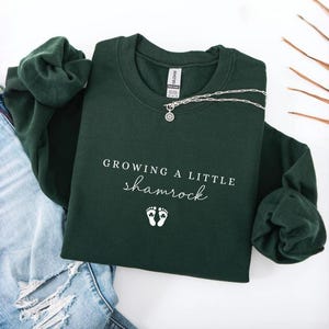 May include: A forest green sweatshirt with the text "Growing a little shamrock" and two small foot prints. A silver necklace with a circular pendant is draped across the sweatshirt. The sweatshirt is folded on top of a pair of blue jeans.