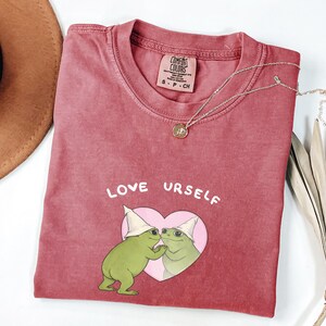 Pu&ograve; includere: T-shirt color cremisi con la scritta "LOVE URSELF" e una grafica di due rane verdi in un cuore rosa. Nel'immagine sono presenti anche una collana d'oro e un cappello marrone.