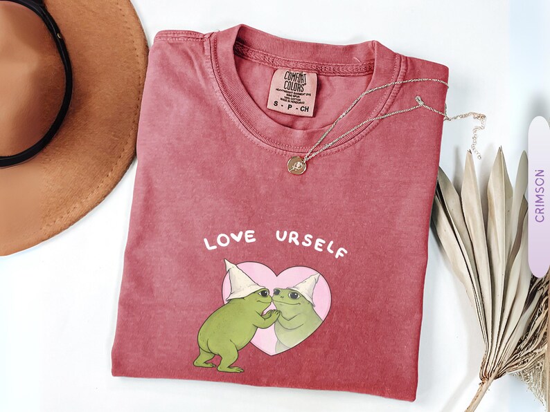 Pu&ograve; includere: T-shirt color cremisi con la scritta "LOVE URSELF" e una grafica di due rane verdi in un cuore rosa. Nel'immagine sono presenti anche una collana d'oro e un cappello marrone.