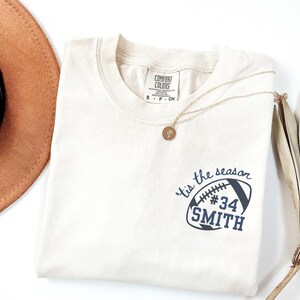 Peut inclure: T-shirt ivoire avec un motif de football bleu marine indiquant "'tis the season #34 SMITH." Un collier doré avec un pendentif est posé sur le t-shirt. Un chapeau marron est visible en haut à gauche.