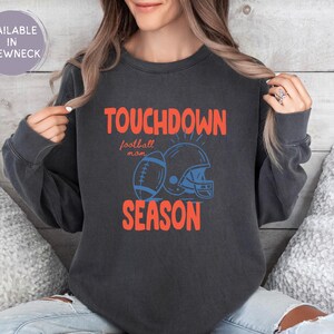 Puede incluir: Sudadera gris oscuro con las palabras "TOUCHDOWN SEASON" en naranja, con un gr&aacute;fico de f&uacute;tbol americano y casco en azul. Las palabras "football mom" est&aacute;n en una fuente m&aacute;s peque&ntilde;a. Disponible en cuello redondo.