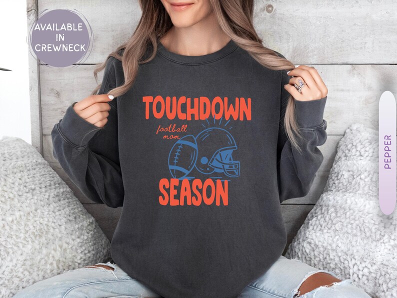Puede incluir: Sudadera gris oscuro con las palabras "TOUCHDOWN SEASON" en naranja, con un gr&aacute;fico de f&uacute;tbol americano y casco en azul. Las palabras "football mom" est&aacute;n en una fuente m&aacute;s peque&ntilde;a. Disponible en cuello redondo.