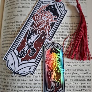 May include: Two decorative bookmarks with a red tassel. The bookmarks feature a stylized illustration of a figure with a sun-like face, holding a sword. One bookmark has a holographic effect.