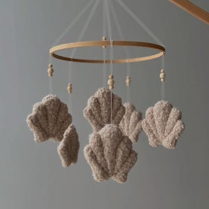 May include: A baby mobile with a wooden ring and hanging beige seashell-shaped ornaments. The mobile is suspended from a wooden arm and includes small wooden beads. The seashells have a textured, plush appearance.