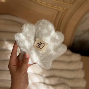 May include: A white, plush, shell-shaped ornament with a soft, fuzzy texture. Centered on the front is a small, rectangular, off-white fabric tag featuring the number 10 written in a simple, dark, handwritten-style font.