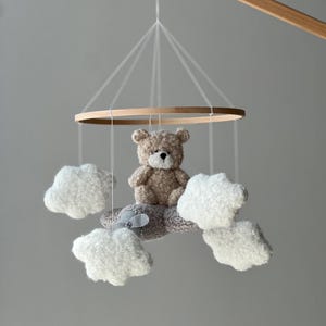 May include: A baby mobile featuring a plush teddy bear and fluffy white clouds suspended from a wooden hoop. The bear is light brown, and the clouds are soft and textured. The mobile is designed for a nursery.