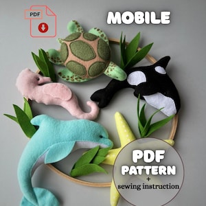 May include: A felt mobile featuring sea creatures: a green turtle, a pink seahorse, a blue dolphin, and a black and white orca. The mobile includes a yellow starfish and green felt seaweed, all attached to a wooden hoop. The text "MOBILE" and "PDF PATTERN + sewing instruction" are also visible.