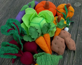 Può includere: Una collezione di cibi in feltro, tra cui un cavolo verde, carote arancioni, barbabietole rosse, una melanzana viola, una patata marrone e zucche arancioni.