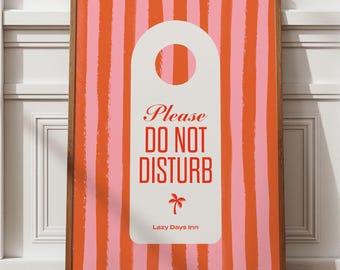May include: A framed wall art print featuring a pink and orange striped background. The artwork includes a white door hanger shape with the words "Please DO NOT DISTURB" in red, and a palm tree graphic. The text "Lazy Days Inn" is also included.