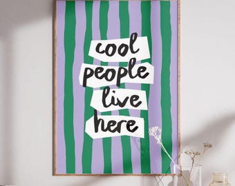 May include: A framed art print with a purple and green striped background. The text "cool people live here" is displayed in a layered, torn-paper style. The artwork is displayed on a wooden surface.