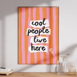 May include: A framed art print with a colorful design. The background features vertical stripes in orange and pink. The words "cool people live here" are written in black on white cut-out shapes.