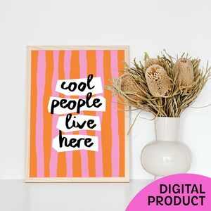 May include: A rectangular framed print featuring vertical orange and pink stripes with the text "cool people live here" in black handwritten letters on white paper strips. Next to the frame is a white vase containing dried brown flowers.
