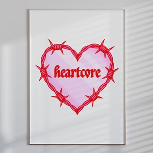 Può includere: Stampa incorniciata con un cuore rosa circondato da filo spinato rosso. La parola "heartcore" è scritta in rosso all'interno del cuore. L'opera è presentata su uno sfondo bianco.