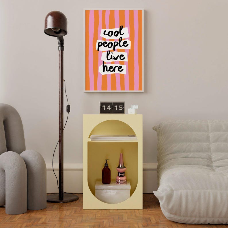 May include: A light yellow rectangular side table with an oval cutout, featuring a shelf holding a dark bottle, a pink decorative object, and a white box. Above it hangs a framed print with orange and pink vertical stripes and the text cool people live here.