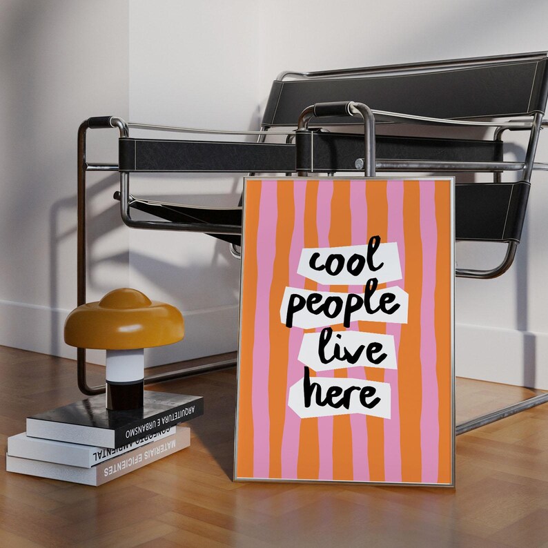May include: A framed art print with a colorful design. The artwork features vertical stripes in pink and orange, with the words "cool people live here" in a bold, modern font. The print is displayed in a room with a chair and a lamp.