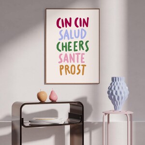 May include: A framed art print with the words "CIN CIN SALUD CHEERS SANTE PROST" in various colours. The print is displayed above a modern black table with decorative candles and a light blue vase on a pink side table.