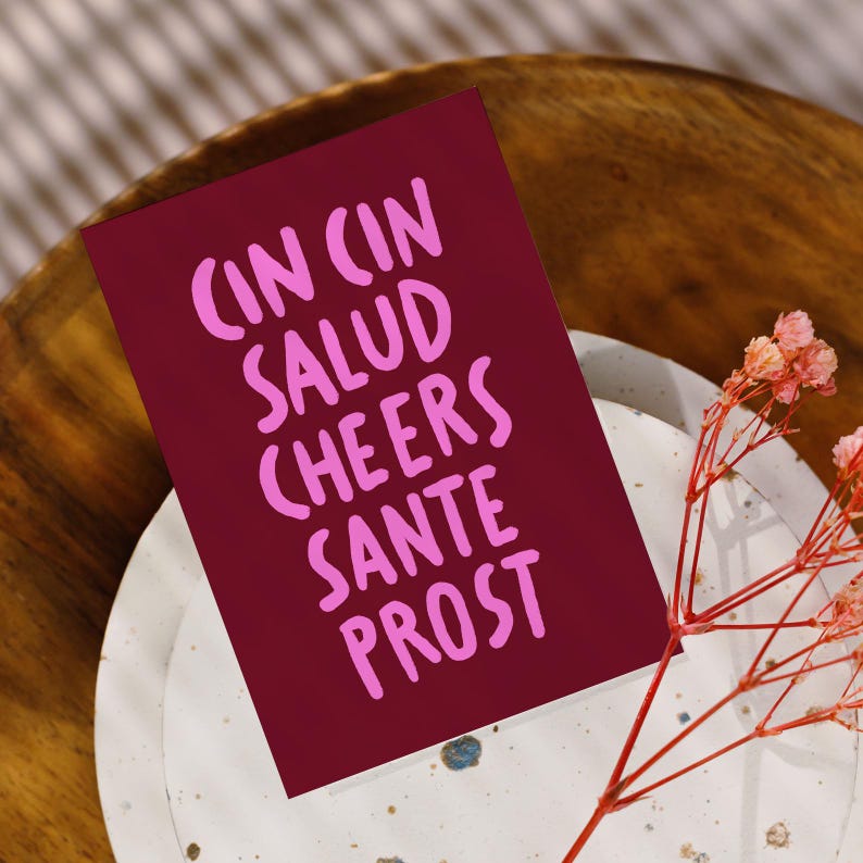 Peut inclure: Une carte bordeaux avec les mots "CIN CIN SALUD CHEERS SANTE PROST" en lettres roses. La carte repose sur une assiette blanche avec un motif tachet&eacute; et un bol en bois. De d&eacute;licates fleurs roses sont au premier plan.