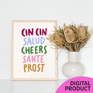 May include: A framed print with the words "CIN CIN SALUD CHEERS SANTE PROST" in various colours. A white vase with dried flowers sits to the right of the print. A pink circle with the words "DIGITAL PRODUCT" is in the lower right corner.