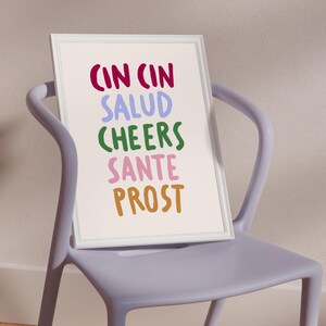 May include: A framed print with the words "CIN CIN SALUD CHEERS SANTE PROST" in various colours. The print is in a white frame and rests on a light purple chair.