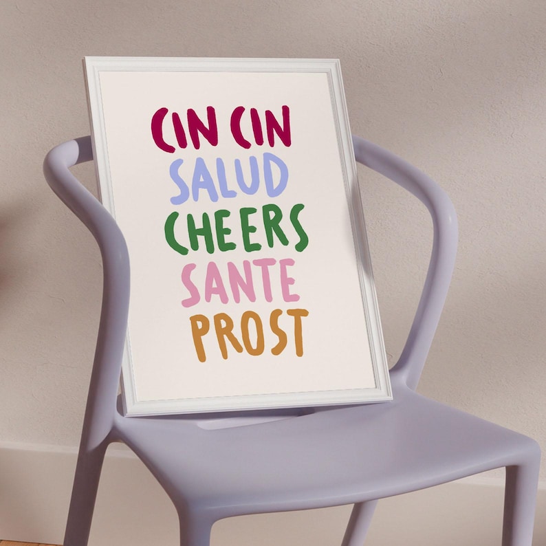 May include: A framed print with the words "CIN CIN SALUD CHEERS SANTE PROST" in various colours. The print is in a white frame and rests on a light purple chair.