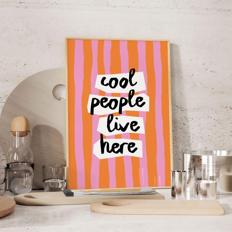 May include: A framed art print with a vibrant orange and pink striped background. The text "cool people live here" is displayed in black, block letters on white cut-out shapes. The artwork is displayed on a white surface.