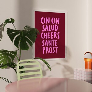 Peut inclure: Un cadre avec un fond bordeaux et un texte rose qui dit "CIN CIN SALUD CHEERS SANTE PROST". L'impression est dans un cadre blanc et est accroch&eacute;e &agrave; un mur beige. Une plante verte et une chaise vert clair sont au premier plan.
