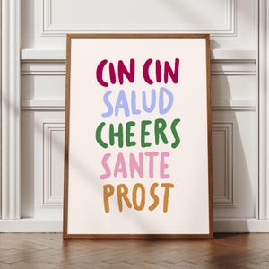 May include: A framed art print featuring the words "CIN CIN SALUD CHEERS SANTE PROST" in a variety of colours. The text is in a playful, handwritten style. The frame is a warm brown, and the background is a light cream colour.