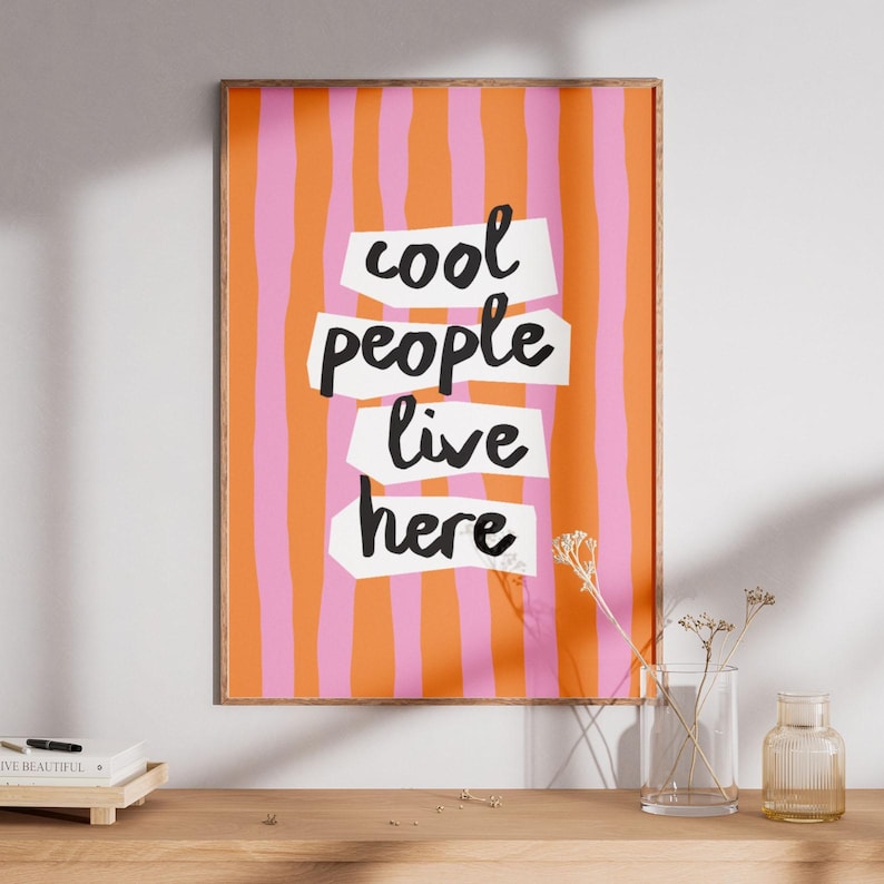 May include: A framed art print with a colorful design. The background features vertical stripes in orange and pink. The words "cool people live here" are written in black on white cut-out shapes.