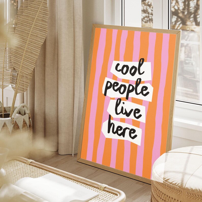 May include: A framed art print with a vertical striped background in orange and pink. The text "cool people live here" is displayed in a bold, handwritten style. The frame is made of light-colored wood.