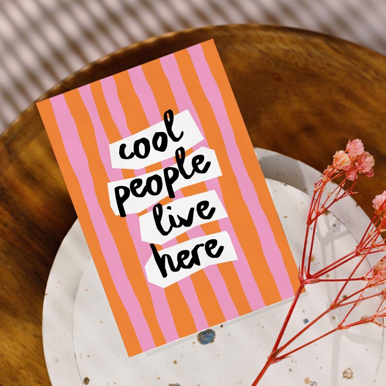 May include: Greeting card with a pink and orange striped background. The words "cool people live here" are in a bold, handwritten style. The card is on a white plate with a wooden bowl in the background. A sprig of pink flowers is in the foreground.