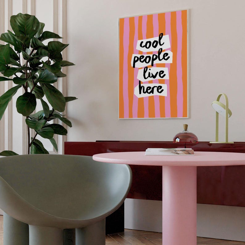 May include: A framed art print with a colorful design. The artwork features vertical stripes in orange and pink, with the words "cool people live here" in a bold, black font. The print is displayed in a modern interior setting.
