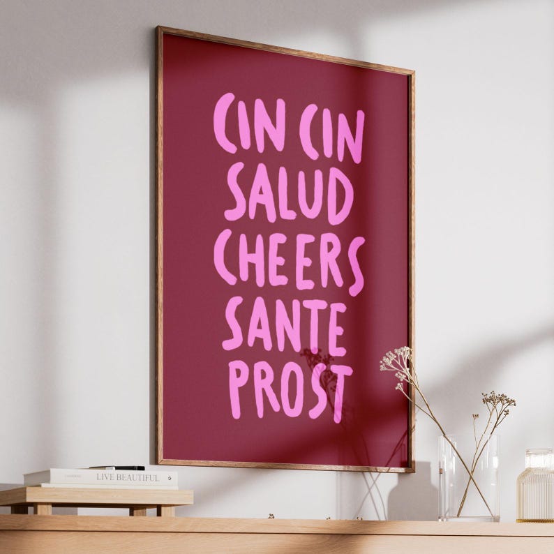 Peut inclure: Une affiche encadr&eacute;e avec un fond bordeaux et du texte rose. Le texte comprend les mots "CIN CIN, SALUD, CHEERS, SANTE, PROST". Le cadre est de couleur marron chaud. L'affiche est expos&eacute;e sur une &eacute;tag&egrave;re en bois.